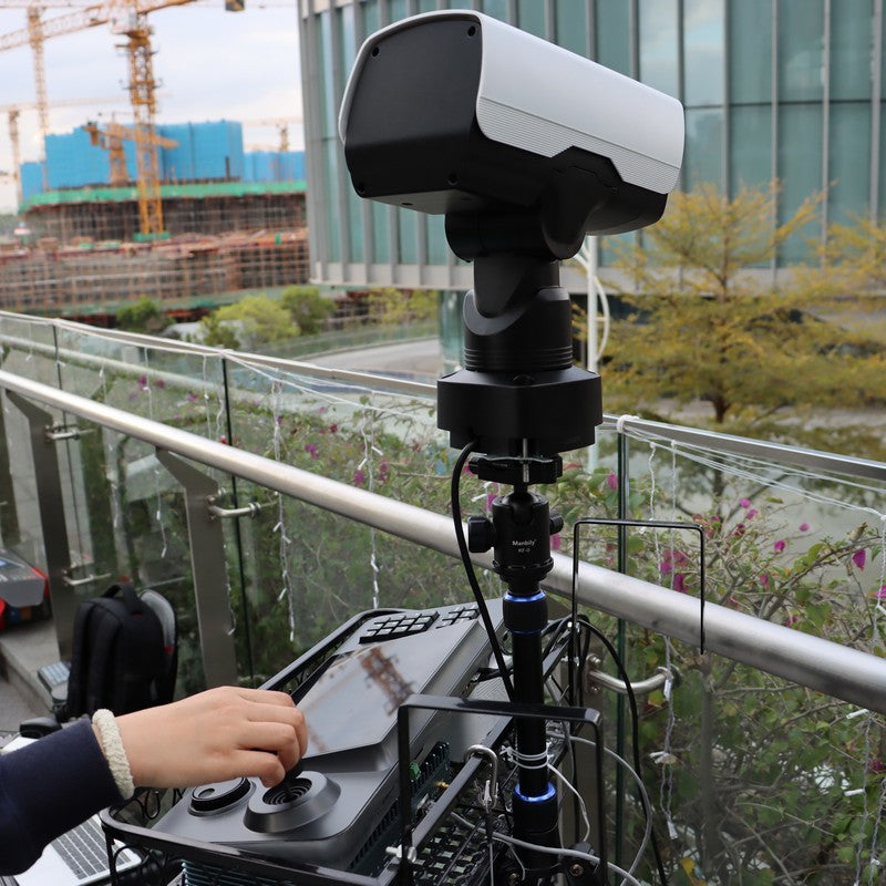 Camera on a tripod with a person adjusting settings, outdoor setting with buildings and plants in the background